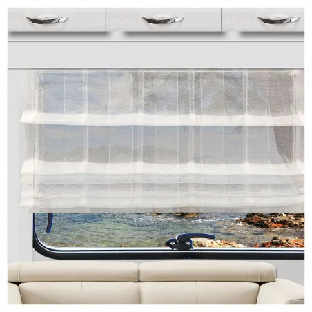 Ein durchsichtiger weißer Vorhang verdeckt teilweise ein großes Fenster mit Blick auf eine felsige Küste und ruhiges blaues Wasser. Über dem Fenster sind weiße Schränke mit silbernen Griffen angebracht, während sich darunter ein beiges Sofa befindet.