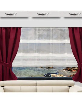 Ein Fenster mit weißen Gardinen und weinroten Vorhängen gibt den Blick auf eine felsige Meeresküste frei. Im Vordergrund befindet sich ein cremefarbenes Sofa. Über dem Fenster sind weiße Schränke mit Chromgriffen angebracht.