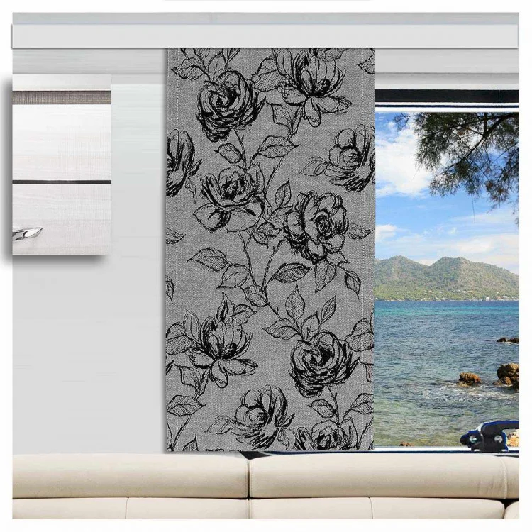 Ein Wohnwagenfenster ist teilweise durch einen grauen Vorhang mit schwarzem Blumenmuster verdeckt. Der unbedeckte Teil gibt den Blick auf das Meer, die Hügel und die Bäume draußen frei. Im Vordergrund steht eine weiße Couch, und links ist ein Wandschrank zu sehen.
