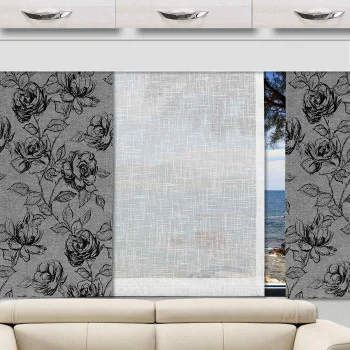 Ein beiges Sofa steht vor einem Wohnwagenfenster mit drei Paneelen - zwei graue Paneele mit schwarzen Blumenmustern auf beiden Seiten und ein durchsichtiges weißes Paneel in der Mitte. Außerhalb des Fensters sind das blaue Meer und der blaue Himmel sowie die Zweige der Bäume zu sehen.