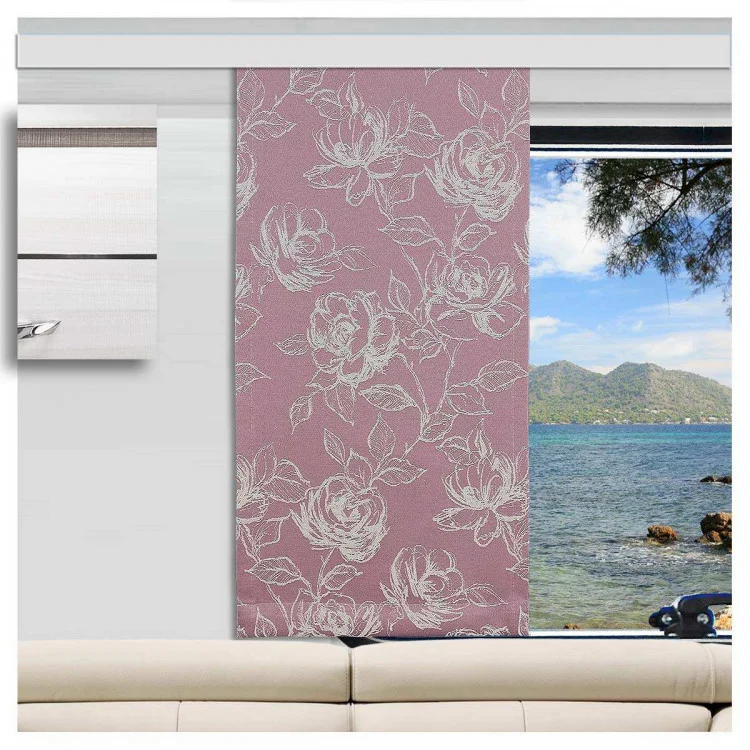 Ein Caravan mit einem hellbeigen Sofa vor einem Fenster. Das Fenster hat einen rosafarbenen, blumengemusterten Vorhang, der teilweise zur Seite gezogen ist und den Blick auf ein blaues Meer, eine felsige Küste, grüne Bäume und ferne Berge unter einem klaren Himmel freigibt.