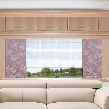 Ein beigefarbenes Sofa steht vor einem Caravanfenster mit weißen Vorhängen und rosafarbenen, blumengeschmückten Seitenwänden. Über dem Fenster sind helle Holzschränke mit silbernen Griffen angebracht. Der Blick nach draußen zeigt eine grüne Landschaft mit Bäumen.