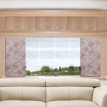 Ein beigefarbenes, gepolstertes Sofa steht vor einem Fenster mit weißen Vorhängen und rosafarbenen, blumengemusterten Seitenwänden. Über dem Fenster stehen drei helle Holzschränke mit silbernen Griffen. Der Blick nach draußen zeigt grüne Bäume und einen blauen Himmel.