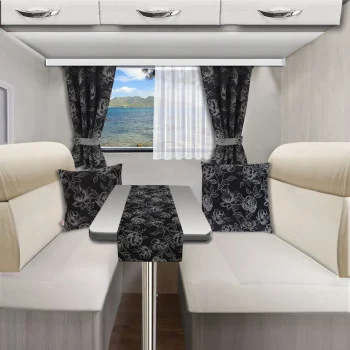 Essbereich im Wohnmobil mit cremefarbenen gepolsterten Sitzen, einem rechteckigen Tisch mit schwarzem Blumenläufer, passenden schwarzen Blumenkissen und Vorhängen sowie einem Fenster, das den Blick auf einen See und Bäume freigibt.