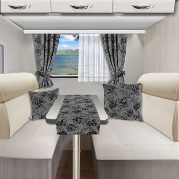 Ein Wohnmobil-Essbereich mit cremefarbenen, gepolsterten Bänken, einem Tisch und passenden, blumengemusterten Kissen, Vorhängen und Tischläufer. Ein Heckfenster zeigt den Blick auf einen See und die Berge bei teilweise bewölktem Himmel. Oberhalb des Fensters befinden sich Hängeschränke.