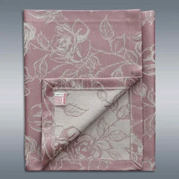 Eine gefaltete Tischdecke mit einem rosa und beigen Blumenmuster ist vor einem grauen Hintergrund mit Farbverlauf angeordnet. Eine Ecke ist umgedreht, um die Rückseite zu zeigen, auf der dasselbe Blumenmuster in umgekehrten Farben zu sehen ist. Auf der Rückseite ist ein kleines Stoffetikett zu sehen.