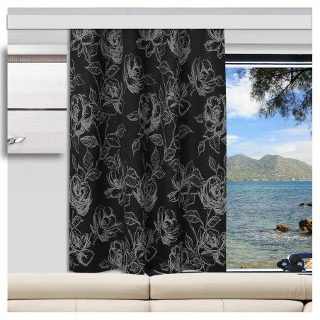 Ein schwarzer Vorhang mit einem weißen Rosenmuster ist teilweise vor ein Fenster gezogen. Aus dem Fenster blickt man auf blaues Wasser, eine felsige Küste, Baumzweige und ferne grüne Berge. Direkt unter dem Fenster steht eine helle Couch.
