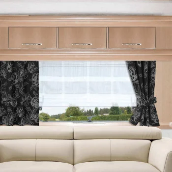 Ein beiges Sofa steht in einem hellen, holzgetäfelten Wohnwagen vor einem Fenster mit weißen Gardinen und schwarzen, blumengemusterten Vorhängen. Darüber sind drei Holzschränke mit silbernen Griffen angebracht. Durch das Fenster sind grüne Bäume und der blaue Himmel zu sehen.