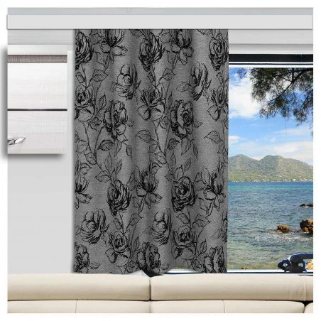 Ein grauer Vorhang mit einem schwarzen Blumenmuster ist vor einem Wohnwagenfenster teilweise geöffnet. Durch das Fenster sind blaues Wasser, ein felsiges Ufer, ferne grüne Hügel und ein Baumzweig zu sehen. Direkt unter dem Fenster steht ein beiges Sofa.