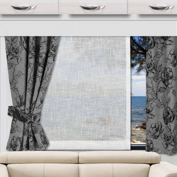 Ein Wohnwagenfenster ist mit weißen Vorhängen und zwei grauen Vorhängen mit Blumenmuster versehen, von denen einer zurückgebunden und einer aufgehängt ist. Darüber sind weiße Schränke mit silbernen Griffen angebracht. Draußen sind das blaue Meer, der Himmel und ein Baumzweig zu sehen.