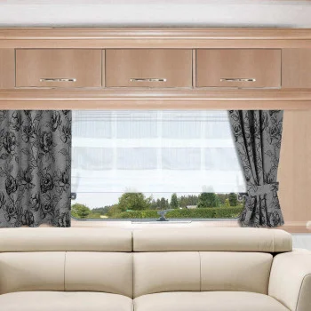 Ein beiges Sofa steht vor einem großen Fenster, das mit weißen Vorhängen und grauen, floral gemusterten Seitenvorhängen versehen ist. Drei helle Holzschränke mit silbernen Griffen sind über dem Fenster angebracht. Draußen sind Bäume und Grünpflanzen zu sehen.