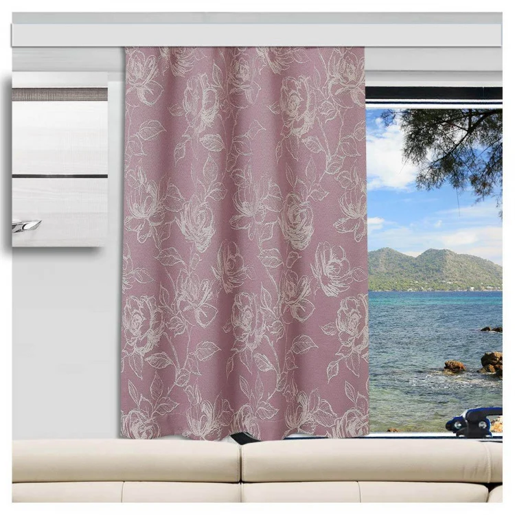 In einem Wohnwagen steht ein beiges Sofa vor einem großen Fenster. Das Fenster bietet einen Blick auf den Ozean mit blauem Wasser, einer felsigen Küste, grünen Hügeln und einem Teil eines Baumzweigs. Ein rosafarbener Vorhang mit einem weißen Blumenmuster verdeckt das Fenster teilweise.