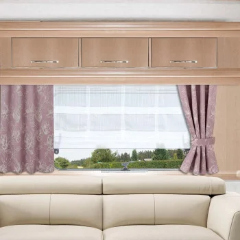 Ein beigefarbenes Sofa steht vor einem Fenster mit rosafarbenen, geblümten Vorhängen, die an den Seiten zugezogen sind und eine Szene im Freien mit Bäumen, Gras und Himmel zeigen. Über dem Fenster sind hölzerne Schränke mit silbernen Griffen angebracht.