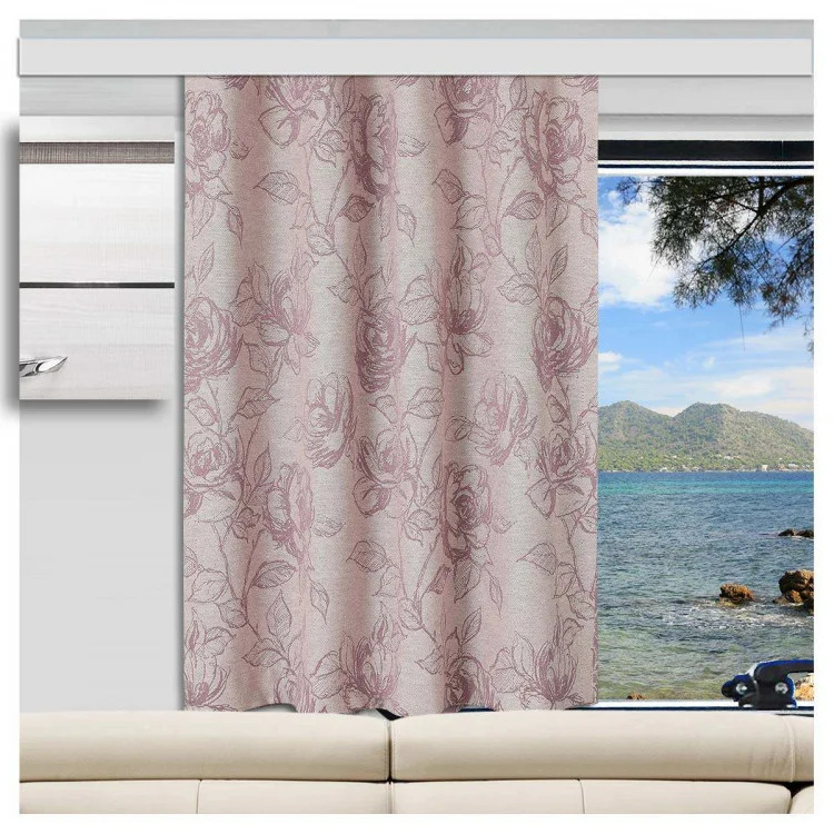 In einem Wohnwagen steht ein beiges Sofa vor einem großen Fenster. Die rosafarbenen, blumengemusterten Vorhänge sind zur Seite gezogen und verdecken teilweise den Blick auf das blaue Meer, die felsige Küste und die grünen Berge unter dem klaren Himmel.