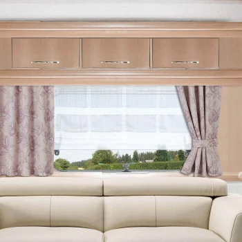 Ein beiges Sofa steht vor einem Fenster, an dem weiße Gardinen und gemusterte rosafarbene Vorhänge befestigt sind. Über dem Fenster sind drei Holzschränke mit silbernen Griffen angebracht. Aus dem Fenster blickt man auf grüne Bäume und einen blauen Himmel.