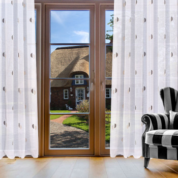 Große Holzfenster in einem Wohnzimmer, dekoriert mit halbtransparenten Vorhängen mit Punkt-Muster. Davor steht ein Sessel.