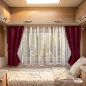 Das Innere eines Wohnwagens besteht aus einem ordentlich gemachten Bett, das unter einem großen Fenster steht, das mit weißen Gardinen und tiefroten Vorhängen verhängt ist. An den Wänden über dem Bett sind Holzschränke angebracht, und natürliches Licht fällt in den Raum und erhellt ihn.