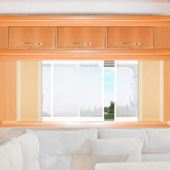 Ein Holzschrank mit drei Schubladen ist über einem großen Fenster angebracht. Das Fenster ist mit weißen Vorhängen verkleidet und gibt den Blick auf das Grün und den blauen Himmel frei. Unter dem Fenster befindet sich ein weißes, gepolstertes Sofa mit mehreren Kissen.