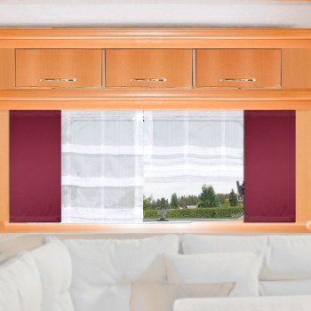 Ein Fenster mit durchsichtigen weißen Vorhängen und weinroten Seitenteilen ist in eine helle Holzwand eingelassen, über der drei kleine Schränke stehen. Durch das Fenster sind Grünpflanzen und Bäume zu sehen. Der untere Teil eines hellen Sofas erscheint am unteren Rand des Bildes.