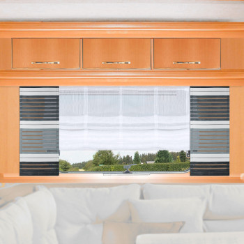 Ein großes Fenster mit weißen Vorhängen und schwarz-grau gestreiften Jalousien ist in eine helle Holzwand über einem weißen gepolsterten Sofa eingelassen. Draußen gibt es Bäume und Grün unter einem bewölkten Himmel. Über dem Fenster sind drei Holzschränke angebracht.