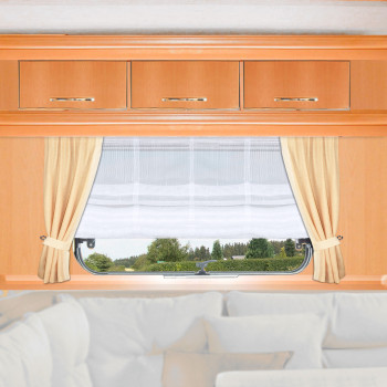 Ein Fenster mit weißen Gardinen und beigen Vorhängen ist in eine Holzwand eingelassen, über der drei Schränke stehen. Durch das Fenster sind grüne Bäume und Sträucher unter einem bewölkten Himmel zu sehen. Unter dem Fenster befindet sich eine weiße gepolsterte Sitzecke.