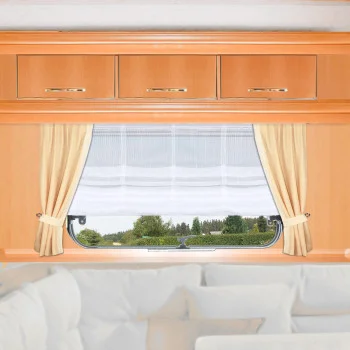 Ein Fenster mit weißen Gardinen und beigen Vorhängen ist in eine Holzwand eingelassen, über der drei Schränke stehen. Durch das Fenster sind grüne Bäume und Sträucher unter einem bewölkten Himmel zu sehen. Unter dem Fenster befindet sich eine weiße gepolsterte Sitzecke.