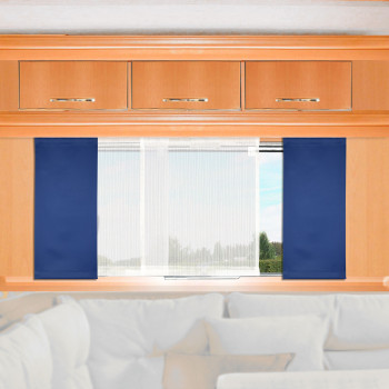 Ein heller Holzschrank mit drei Schubladen steht über einem Fenster, das von blauen Seitenvorhängen und einem durchsichtigen weißen Mittelvorhang eingerahmt wird. Draußen sind Bäume und der Himmel zu sehen. Eine weiße Couch mit beigen Kissen ist teilweise unter dem Fenster zu sehen.