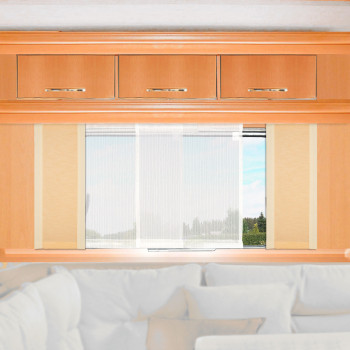 Ein hölzerner Schrank mit drei Schubladen ist über einem Fenster angebracht, das von weißen Vorhängen verdeckt wird. Durch das Fenster sieht man Grün und einen blauen Himmel. Unter dem Fenster steht eine weiße Couch mit cremefarbenen Kissen. Der Raum ist gut beleuchtet.