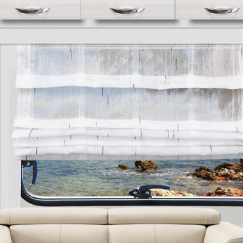 Das weiße Raffrollo Alva ist teilweise über ein Fenster gezogen, das den Blick auf das blaue Wasser und eine felsige Küste freigibt. Unter dem Fenster steht ein beiges Sofa, darüber befinden sich weiße Schränke mit Metallgriffen. Der Raum wird von Tageslicht hell erhellt.