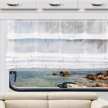 Das weiße Raffrollo Alva ist teilweise über ein Fenster gezogen, das den Blick auf das blaue Wasser und eine felsige Küste freigibt. Unter dem Fenster steht ein beiges Sofa, darüber befinden sich weiße Schränke mit Metallgriffen. Der Raum wird von Tageslicht hell erhellt.