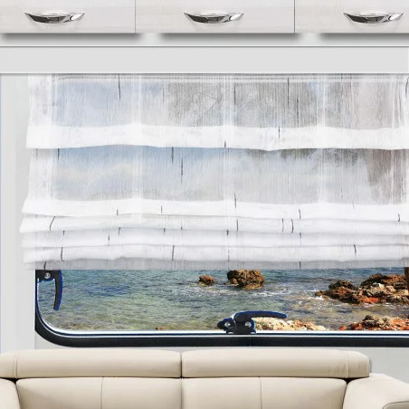 Das weiße Raffrollo Alva ist teilweise über ein Fenster gezogen, das den Blick auf das blaue Wasser und eine felsige Küste freigibt. Unter dem Fenster steht ein beiges Sofa, darüber befinden sich weiße Schränke mit Metallgriffen. Der Raum wird von Tageslicht hell erhellt.