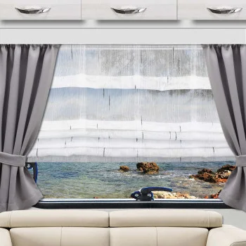 Ein Fenster mit grauen Vorhängen und weißen, durchsichtigen Jalousien bietet einen Blick auf eine felsige Küste und klares, blaues Wasser. Die Ansicht zeigt den Blick aus einem Raum, oberhalb eines beigen Sofas. Über dem Fenster sind weiße Schränke mit silbernen Griffen angebracht.