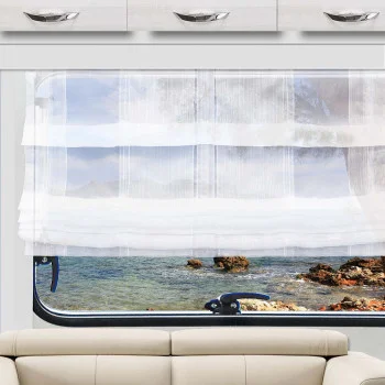 Ein weißer, durchsichtiger Vorhang hängt teilweise über einem Fenster und gibt den Blick auf eine felsige Küste und das blaue Wasser draußen frei. Unterhalb des Fensters steht ein hellbeiges Sofa, darüber befinden sich weiße Schränke mit silbernen Griffen. Der Raum besticht durch neutrale Farbtöne und den Blick auf die