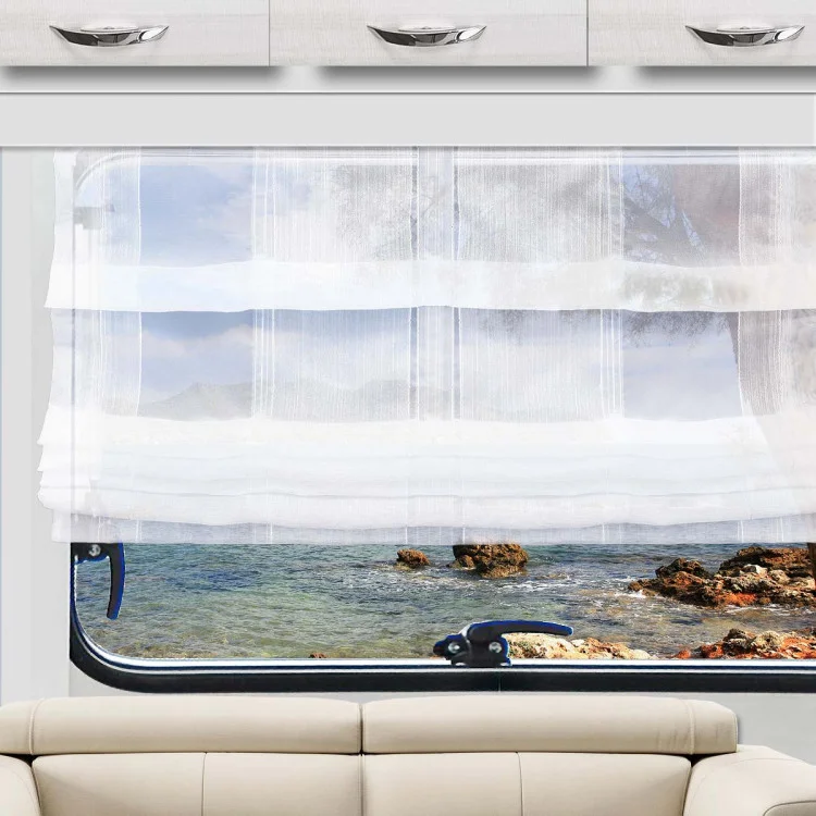 Ein weißer, durchsichtiger Vorhang hängt teilweise über einem Fenster und gibt den Blick auf eine felsige Küste und das blaue Wasser draußen frei. Unterhalb des Fensters steht ein hellbeiges Sofa, darüber befinden sich weiße Schränke mit silbernen Griffen. Der Raum besticht durch neutrale Farbtöne und den Blick auf die