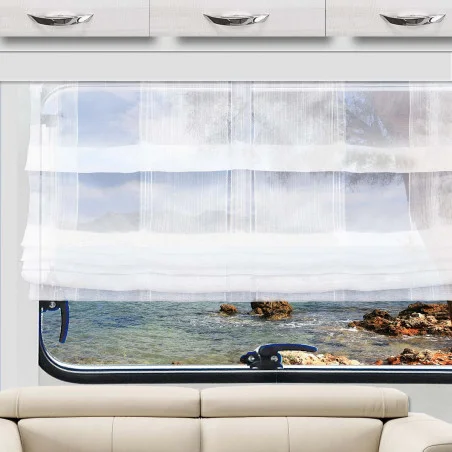 Ein weißer, durchsichtiger Vorhang hängt teilweise über einem Fenster und gibt den Blick auf eine felsige Küste und das blaue Wasser draußen frei. Unterhalb des Fensters steht ein hellbeiges Sofa, darüber befinden sich weiße Schränke mit silbernen Griffen. Der Raum besticht durch neutrale Farbtöne und den Blick auf die