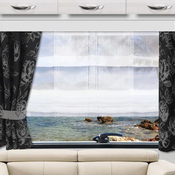 Im Wohnzimmer befindet sich ein Fenster, das mit schwarzen Blumenvorhängen und weißen Gardinen verziert ist und den Blick auf eine felsige Küste und klares blaues Wasser freigibt. Über dem Fenster sind weiße Schränke mit silbernen Griffen angebracht, darunter steht ein beiges Sofa.