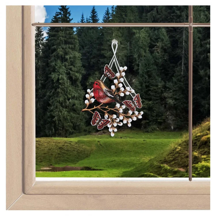 Zarte Vögel und Schmetterlinge aus Spitze schmücken eine bezaubernde Frühlings- und Oster-Schaufensterdekoration vor einer malerischen Kulisse aus grasbewachsenen Kiefern.