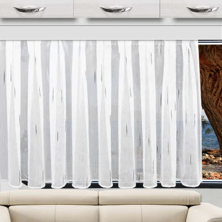 Über einem beigen Sofa hängt ein transparenter Scherli-Vorhang aus der „Caravanstore Alva“-Kollektion von SeGaTeX; durch das Fenster sind Bäume zu sehen, und über dem Sofa sind Schubladen angebracht.