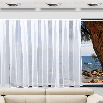 Der Sichtschutz „Lykke Sablé“ von Caravanstore aus der Home-Fashion-Kollektion von SeGaTeX hängt vor einem Fenster mit Blick auf die Küste, daneben stehen ein beiges Sofa und weiße Schränke.