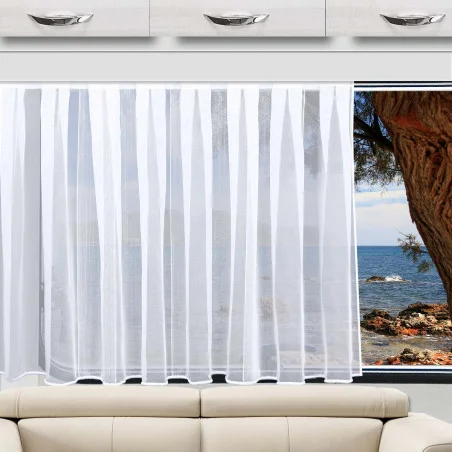 Der Sichtschutz „Lykke Sablé“ von Caravanstore aus der Home-Fashion-Kollektion von SeGaTeX hängt vor einem Fenster mit Blick auf die Küste, daneben stehen ein beiges Sofa und weiße Schränke.