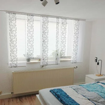 Ein helles Schlafzimmer mit durchsichtigen, gemusterten weißen Vorhängen vor einem breiten Fenster. Auf der Fensterbank stehen Zimmerpflanzen, unter dem Fenster befindet sich ein Heizkörper und ein ordentlich gemachtes Bett mit einer blau-weißen Bettdecke.