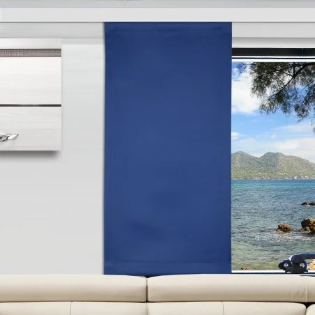 Ein modernes Wohnzimmer mit einem beigen Sofa und einem blauen Flächenvorhang, der ein Fenster teilweise verdeckt. Das Fenster bietet einen Blick auf ein ruhiges Meer, grüne Berge in der Ferne und einen klaren Himmel. Links neben dem Fenster befindet sich ein gerahmtes Wandkunstwerk.