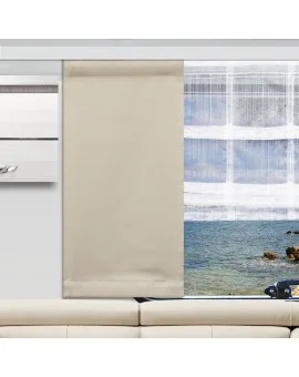 Ein beige Flächenvorhang verdeckt teilweise ein großes Fenster, das sich hinter einem weißen Sofa befindet. Der freiliegende Teil des Fensters gibt den Blick auf das Meer und die Felsen draußen frei. Weiße transparente Vorhänge sorgen für Struktur und auf der linken Seite befindet sich ein Hängeschrank