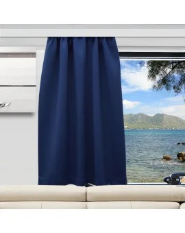 Ein dunkelblauer Vorhang verdeckt die Hälfte eines großen Fensters in einem modernen Wohnwagen. Durch das Fenster blickt man auf einen ruhigen blauen Ozean, Baumzweige und entfernte grüne Hügel unter einem teilweise bewölkten Himmel. Unter dem Fenster befindet sich ein beiges Sofa.
