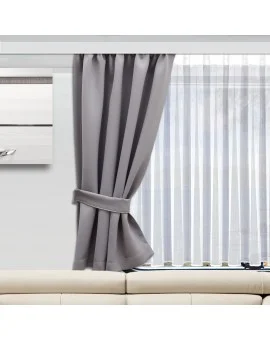 In einem modernen Wohnwagen steht ein beigefarbenes Sofa vor einem großen Fenster mit weißen Vorhängen und einem einzelnen grauen Verdunkelungsvorhang, der mit einem passenden Raffhalter zur Seite gezogen wurde. An der linken Wand ist ein minimalistisches Kunstwerk angebracht.