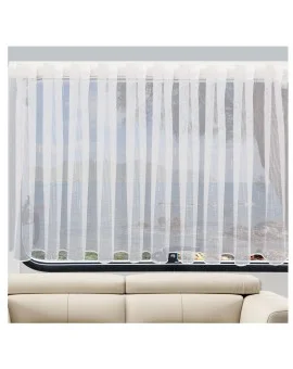 Ein durchsichtiger weißer Vorhang bedeckt ein Fenster und streut das einfallende Licht. Hinter dem Vorhang sind das Meer und eine felsige Küstenlinie zu sehen. Im Vordergrund, unterhalb des Fensters, ist ein Teil eines hellbeigen Sofas zu sehen.