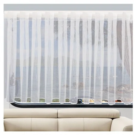 Ein durchsichtiger weißer Vorhang bedeckt ein Fenster und streut das einfallende Licht. Hinter dem Vorhang sind das Meer und eine felsige Küstenlinie zu sehen. Im Vordergrund, unterhalb des Fensters, ist ein Teil eines hellbeigen Sofas zu sehen.