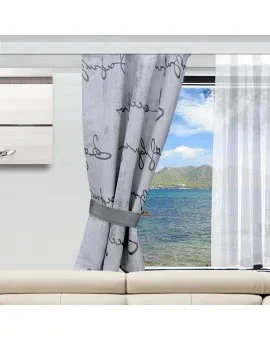In einem modernen Wohnzimmer steht ein beiges Sofa vor einem großen Fenster. Graue Vorhänge mit kursiven Schriftzügen sind zurückgebunden und geben den Blick auf blaues Wasser, grüne Hügel und einen teilweise bewölkten Himmel frei.