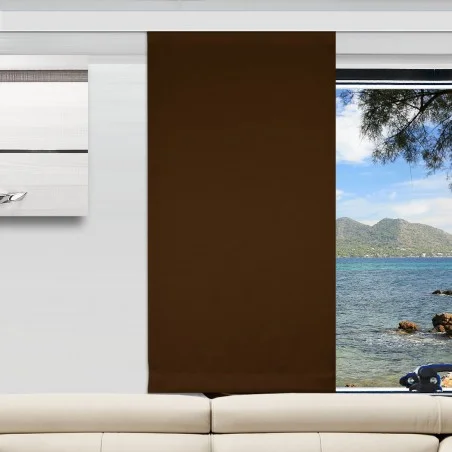 Ein moderner Camperraum mit einer weißen Couch, einem braunen vertikalen Flächenvorhang, der ein Fenster teilweise verdeckt, und Wandkunst auf der linken Seite. Der unbedeckte Teil des Fensters zeigt Berge, blaues Meer und Felsen in der Aussicht nach draußen.